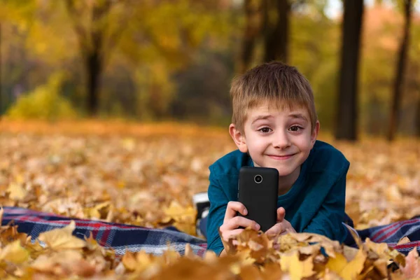 Χαμογελαστός μαθητής με smartphone που βρίσκεται σε καρό, κίτρινα φύλλα του φθινοπώρου. Φθινοπωρινή μέρα — Φωτογραφία Αρχείου