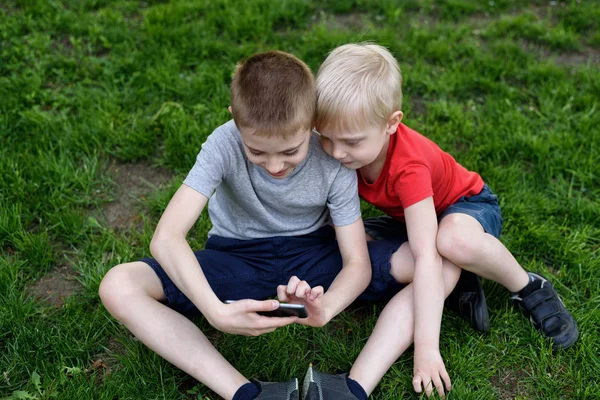 Two boys are gaming on the smartphone while sitting on the grass.