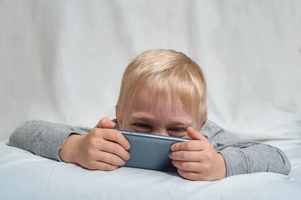 小さなブロンドの男の子は、彼の鼻がベッドに横たわって、スマートフォンに埋もれた何かを見て。ガジェットレジャー — ストック写真