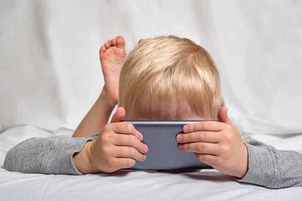Ένα μικρό ξανθό αγόρι που ψάχνει κάτι που η μύτη του θάφτηκε σε ένα smartphone, ξαπλωμένη στο κρεβάτι. Ψυχαγωγία — Φωτογραφία Αρχείου