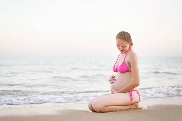 Ung gravid kvinna i baddräkt sitter på stranden och kramar hennes mage. Sommartid före solnedgången Royaltyfria Stockfoton