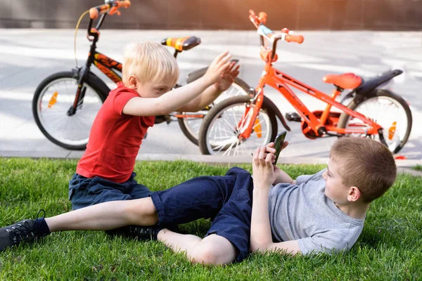 Két fiú fényképezett egy okostelefon ülve a fűben. Pihenés a kerékpározás, a kerékpárok a háttérben — Stock Fotó