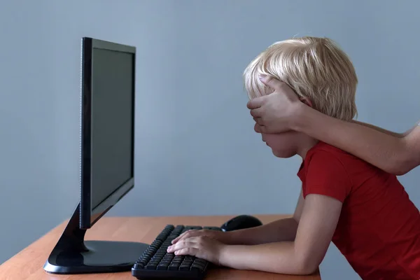 Children hands puts over eyes to a little boy who uses a computer. Dangerous content. Children and gadgets. — Stock Photo, Image