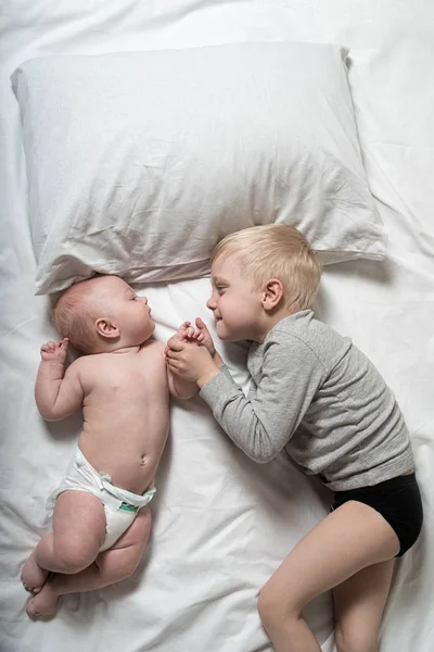 Bebê e sorrindo irmão mais velho estão deitados na cama. Eles brincam, se comunicam e interagem. Vista superior — Fotografia de Stock