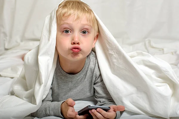 El niño rubio hace muecas con un teléfono inteligente en sus manos. Acostado en la cama y escondido bajo las sábanas. Gadget Ocio —  Fotos de Stock