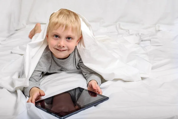 タブレットで微笑む小さなブロンドの男の子。ベッドに横たわって カバーの下に隠れてるガジェットレジャー — ストック写真