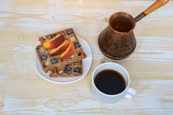 Chocolate Belgian waffles with fruits, cup of coffee and cezve on white wooden background. Delicious breakfast. Top view — Stock Photo, Image