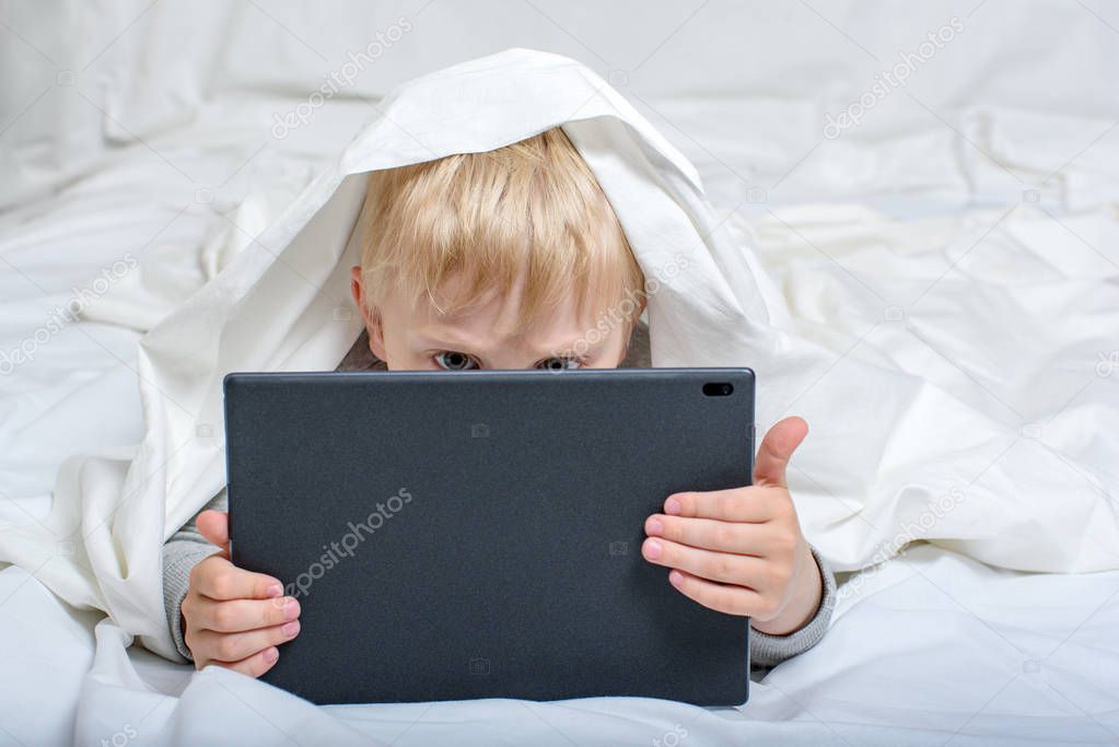 Little blond boy buried his nose in the tablet. Lying in bed and hiding under the covers. Gadget Leisure