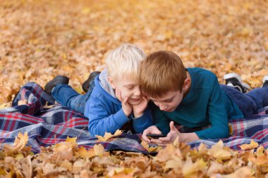 Akıllı telefonu kullanan iki küçük kardeş, sarı sonbahar yapraklarında yatıyorlar. Gülümsüyor ve eğleniyor. Sonbahar günü