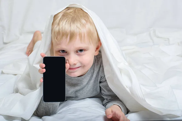 Χαμογελαστό ξανθό αγόρι με έξυπνο τηλέφωνο. Ξαπλωμένη στο κρεβάτι και κρυμμένη κάτω από τα σκεπάσματα. Αναψυχή του Gadget — Φωτογραφία Αρχείου