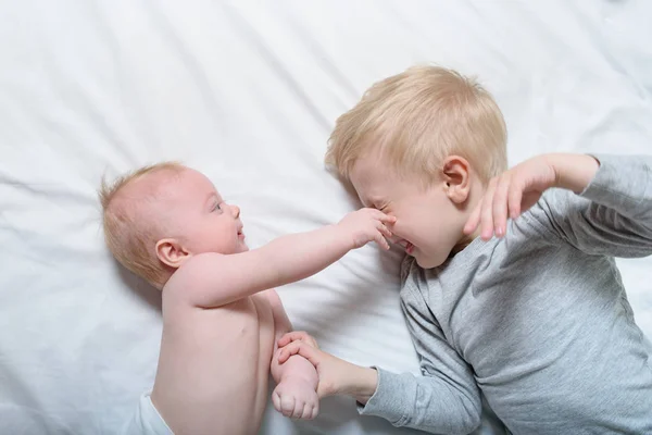 Bebé y hermano mayor sonriente están acostados en la cama. Juegan, son graciosos e interactúan. Vista superior — Foto de Stock