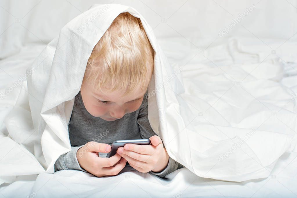 Little blond boy is watching something on a smartphone. Lying in bed and hiding under the covers. Gadget Leisure
