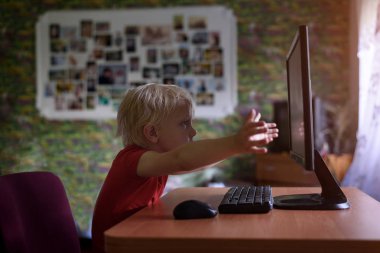 Sarışın çocuk büyük bir bilgisayar monitörünün yanında oturur ve ellerine uzanır..