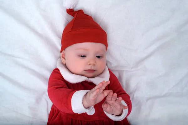 Dziecko w kapeluszu Mikołaja leżące na białym tle, bawiące się rękami. Koncepcja świąteczna. — Zdjęcie stockowe