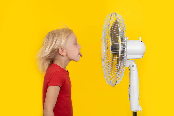 Fair-haired boy shows the tongue vs ventilator. Kid in red shirt on yellow background