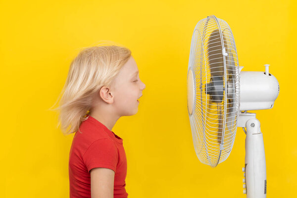 Cute blond child enjoying cold air from fan. Yellow background. Very warm summer.