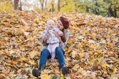 Sonbahar Parkı 'nda düşen yaprakların üzerinde bebeğiyle oturan genç bir anne. Anne ve çocuk dışarıda..