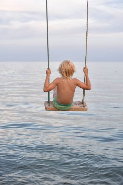Çocuk salıncakta sallanıyor. Denizin çocuklarıyla tatil. Mutlu bir çocukluk. Dikey çerçeve.