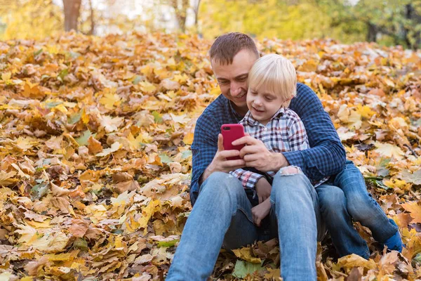 Father and son sitting in autumn Park and looking at phone. Fatherhood.