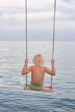 Sarı saçlı çocuk denizde salıncakta sallanıyor. Çocuklarla yaz tatili.