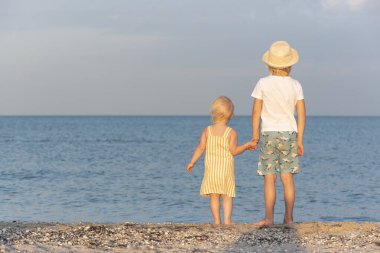 Deniz kenarındaki çocuklar. Arkadan bak. Abisi küçük kız kardeşinin elini tutuyor.