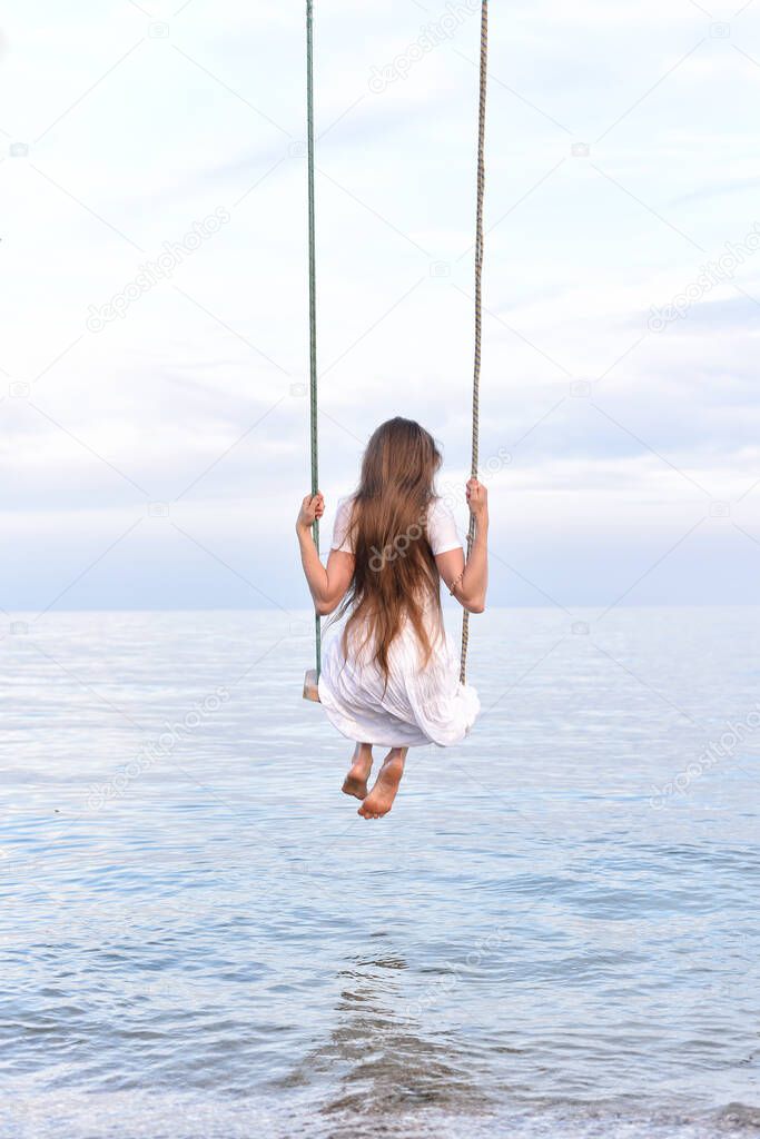 Young girl in white outfit with long hair rides on swing above the water. Romance. Harmony. Vertical frame. Back view.