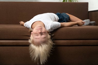 Sarışın çocuk kanepede başı öne eğik yatıyor. Çocuk yorgun. Çocuğun yanlış uyku pozisyonu.