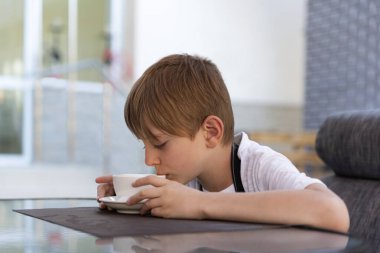 Oğlan kafede çay içiyor. Çocuk beyaz bardaktan kahve içiyor..