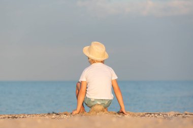 Hasır şapkalı çocuk kumlu sahilde oturuyor ve denize bakıyor. Arka plan.