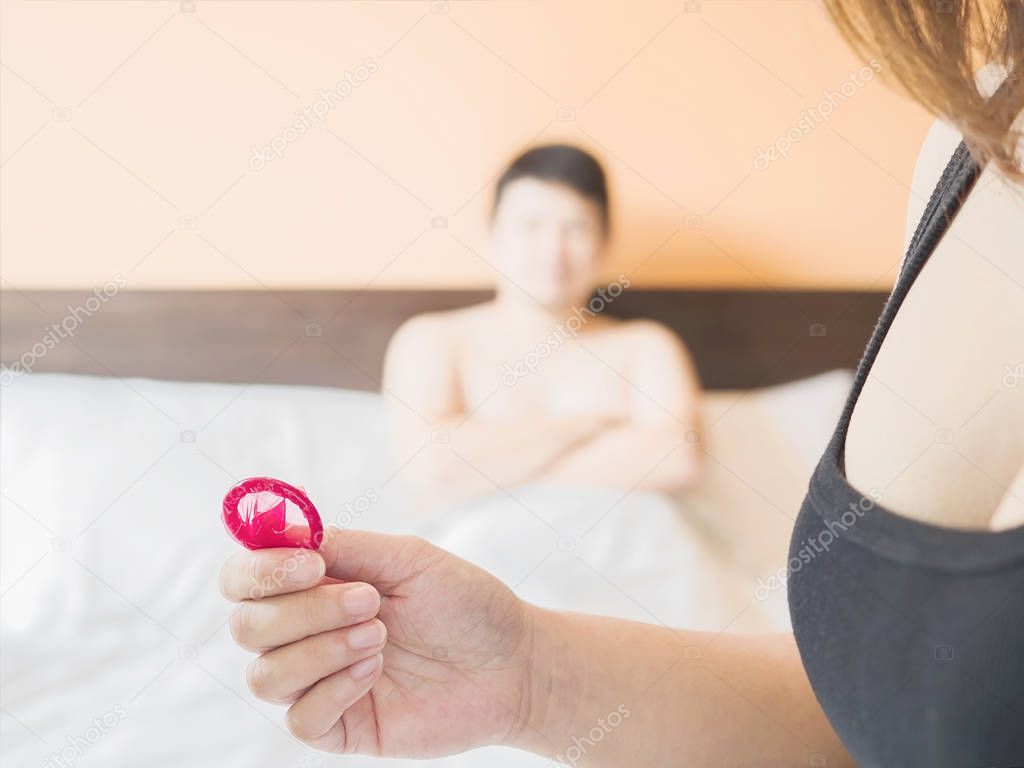 Woman holding red condom with blurred background of a man lie on a bed