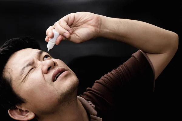 Man dropping eye drop medicine healing his eye pain with black background - health care eye medicine with people concept