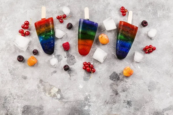 Gökkuşağı renkli dondurma ahududu sarı ve kırmızı, böğürtlen, frenk üzümü ile buzlu şeker ahşap ile gri arka plan üzerinde sopa. Görünümü en çok, uzay kopyalayın. Gıda arka plan