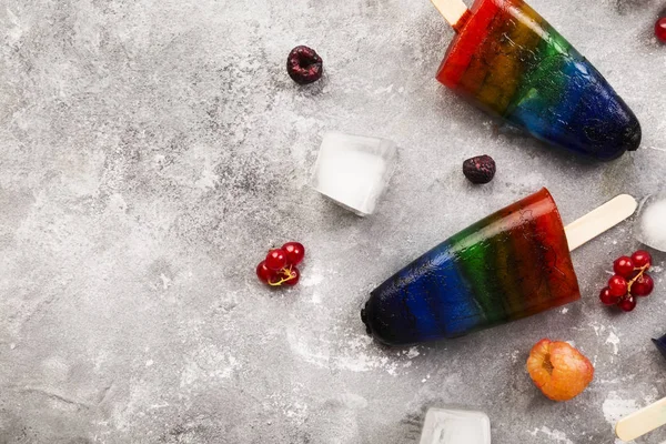 Rainbow colorful ice cream with raspberry yellow and red, blackberry, currant popsicle with wood stick on gray background. Top view, copy space. Food background