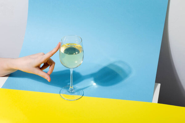 Female hand pushes glass of white wine on yellow blue background, long shadow from the glass. Shadow effect, hard light