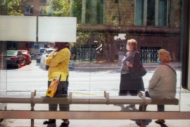 Otobüs durağındaki insanlar tıbbi maskeli, camın arkasından çekim yapıyorlar.