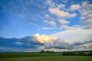 Yeşil tarlaları ve mavi gökyüzünde dramatik bulutları olan bir yaz manzarası. Güneş bulutlardan birini aydınlatıyor, ona sıcak altın bir parıltı veriyor.