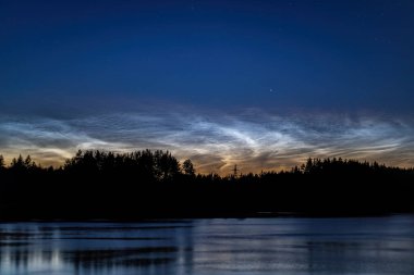 Gökyüzünde parlayan gece parlayan bulutlarla gölün kenarında büyülü bir yaz akşamı. Sakin su mavi alacakaranlık gökyüzünü yansıtır ve karanlık orman silueti ufku çizer.