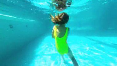 Renkli bir mayo giymiş genç bir kız suyun altında yüzüyor, neşeli hareketler ve akıcılık sergiliyor, güneş ışığı suyun içinden süzülüyor, canlı bir su atmosferi yaratıyor.