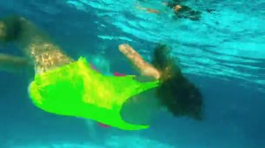 Genç bir kız, parlak bir mayo içinde zarif bir şekilde suyun altında yüzüyor. Etrafı ışıldayan su ve sakin bir su atmosferiyle çevrili akışkan bir hareket ve neşeli bir keşif sergiliyor.