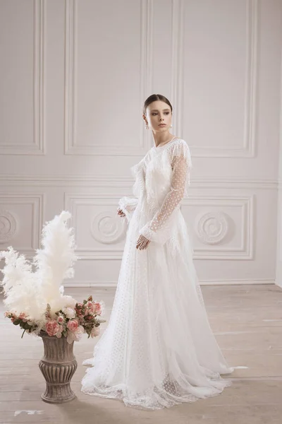 Beyaz, şık, uzun gece elbisesi giymiş, çiçekli vazonun yanında poz veren, stüdyo tüylü sarışın bir kadın.   