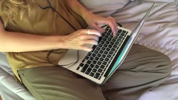 美丽的女人听耳机的音乐和打字笔记本电脑键盘 而坐在床上在家里 — 图库视频影像