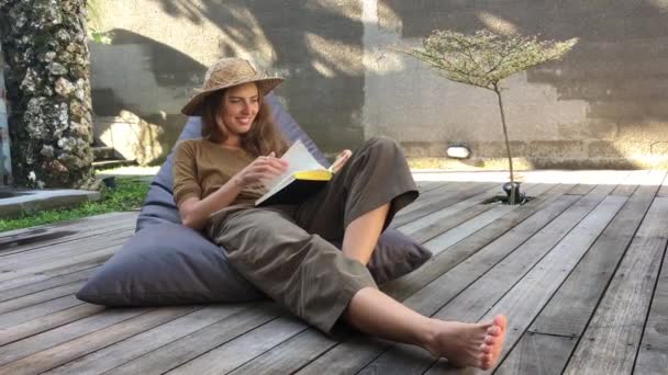 魅力的な女性が本を読み 木製のテラスでビーンバッグの椅子に座りながらリラックス — ストック動画