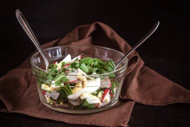 Sebze vejetaryen salata ramson, turp, yeşil soğan ve çatal bıçak takımı peçeteye retro işlemedeki karanlık bir beton zemin üzerine beyaz bir tabak yoğurt sos ile haşlanmış yumurta. Yakın çekim, en iyi görünüm, flatlay. Sağlıklı beslenme kavramı