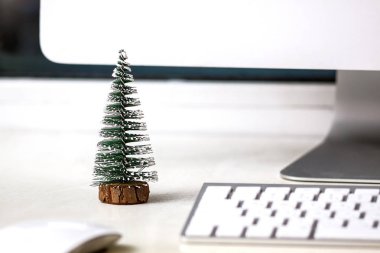 Küçük bir köknar ağacı beyaz bir masaüstünde, bilgisayar monitörü, klavye ve farenin yanında duruyor. Konsept, iş yerinde Noel havası yaratmak.