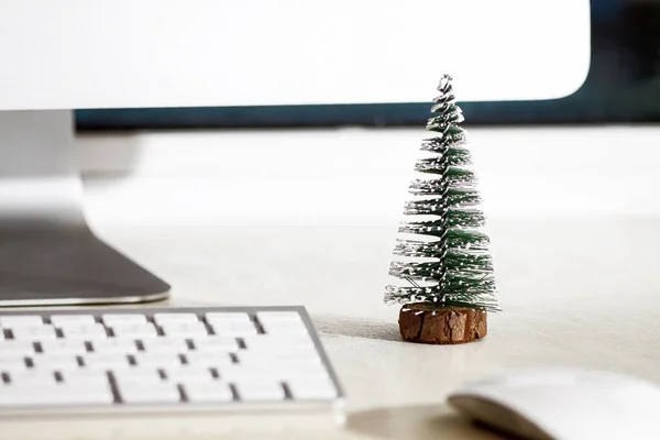 Küçük bir köknar ağacı beyaz bir masaüstünde, bilgisayar monitörü, klavye ve farenin yanında duruyor. Konsept, iş yerinde Noel havası yaratmak.