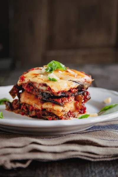 Tradicional PARMIGIANA di Melanzane: pieczone bakłażan-Włochy, sycylijska kuzynka. Pieczone bakłażan z serem, pomidorami i przyprawami na białej płytce. Danie z bakłażan jest na drewnianym stole — Zdjęcie stockowe