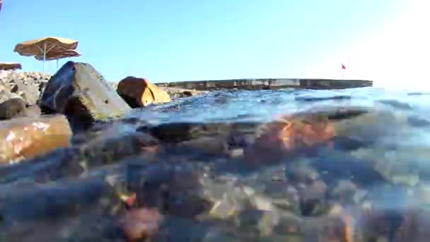 海の波は かわいい傘を持つビーチの沿岸の岩の海岸に転がり あなたは赤い旗 水中ビューを持つ桟橋を見ることができます — ストック動画