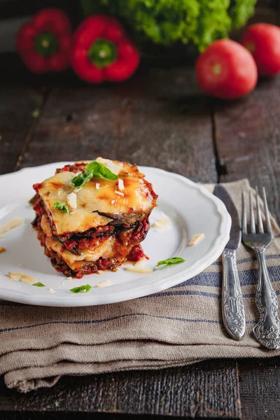 Tradicional PARMIGIANA di Melanzane: pieczone bakłażan-Włochy, sycylijska kuzynka. Pieczone bakłażan z serem, pomidorami i przyprawami na białej płytce. Danie z bakłażan jest na drewnianym stole — Zdjęcie stockowe