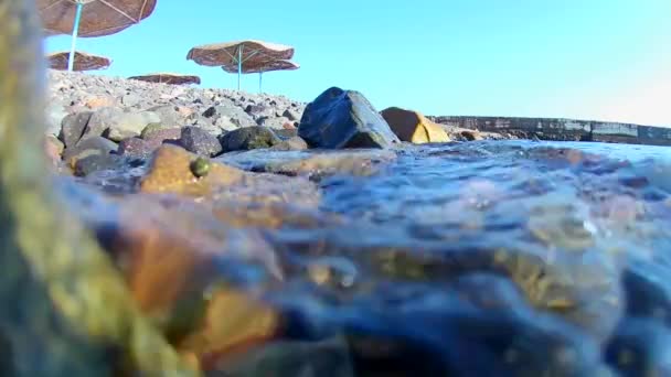 Meereswellen wälzen sich auf die felsige Küste eines Strandes mit reetgedeckten Sonnenschirmen, man sieht einen Pier mit roter Flagge, Unterwasserblick. Zeitlupe — Stockvideo