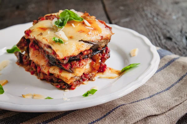 Tradicional Parmigiana di melanzane: berinjela assada - itália, cousina da Sicília. Berinjela assada com queijo, tomates e especiarias em um prato branco. Um prato de berinjela está em uma mesa de madeira — Fotografia de Stock